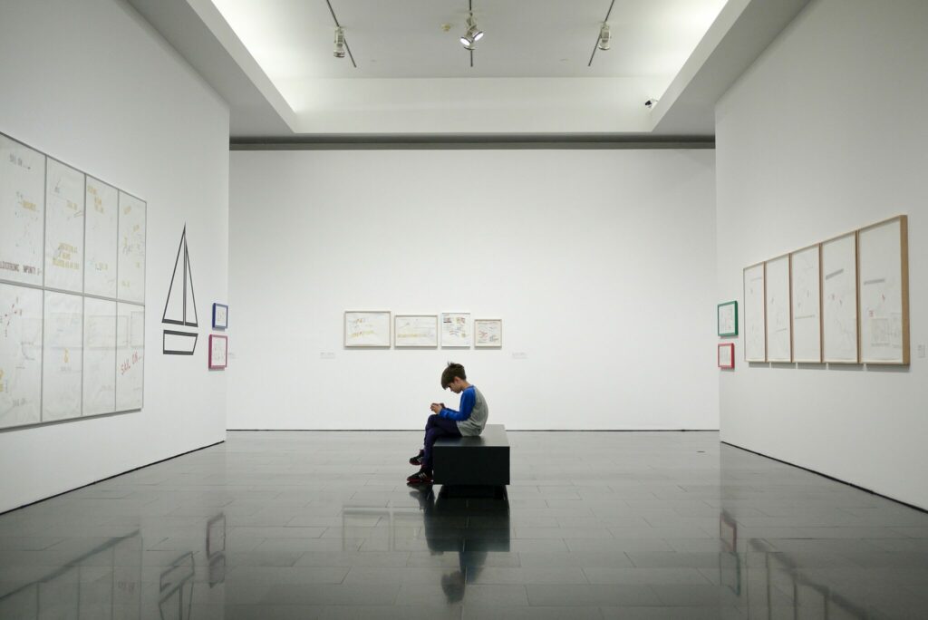 A young person sat in a wide open space at a museum. They are engaged with something in their hands while they sit. They are surrounded by three walls, all with pieces of art work.